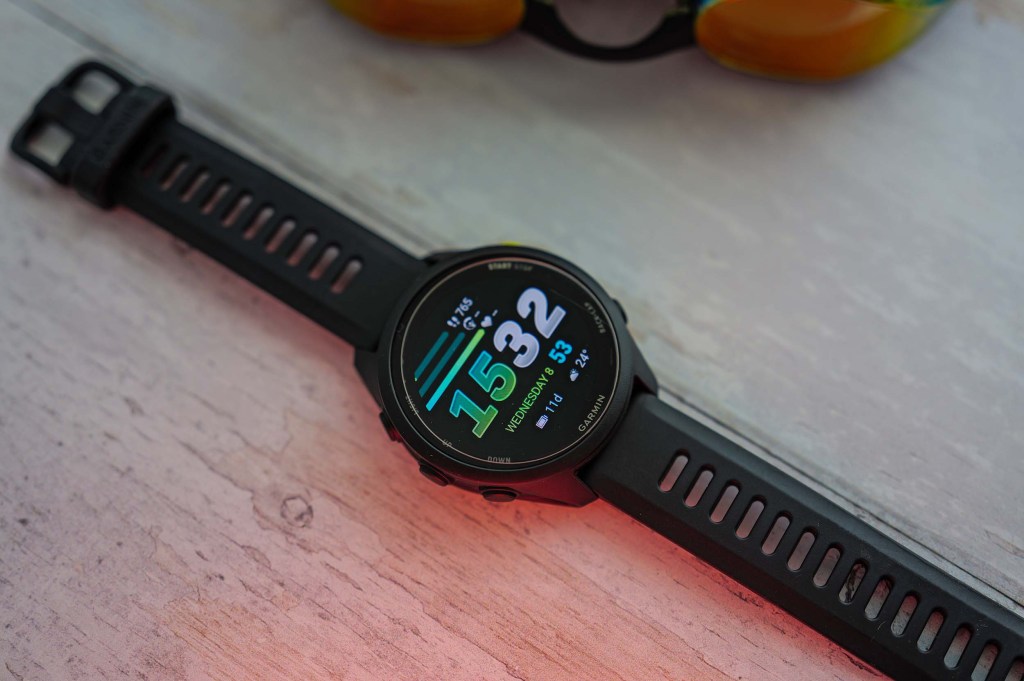 Close-up of a black smartwatch displaying the time, date, and weather information, placed on a light wooden surface next to a pair of sunglasses.