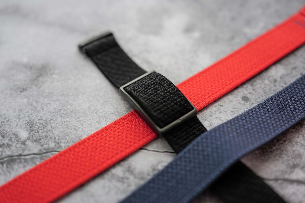 Close-up of three fabric straps: black, red, and navy blue, arranged in a crisscross pattern on a textured surface.